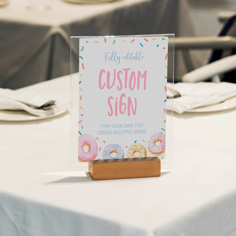 K&ouml;nnte beinhalten: Ein transparenter Acryl-Schildhalter mit einem rechteckigen Holzsockel. Das Schild zeigt einen Rand aus bunten Streuseln, Illustrationen von vier glasierten Donuts und den Text: Fully editable CUSTOM SIGN. TYPE YOUR OWN TEXT CREATE MULTIPLE SIGNS.