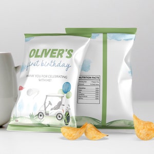 May include: Two white chip bags with a golf theme. The front bag reads "OLIVER'S first birthday" with a golf cart illustration. The back of the bag has a nutrition facts label. A few chips are in front of the bags.