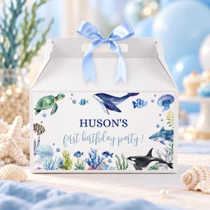 May include: A white gable-style gift box featuring a light blue satin ribbon bow on top. The front displays watercolor illustrations of sea creatures including a turtle, whale, shark, orca, and jellyfish, with the text HUSON'S first birthday party!