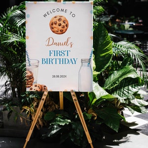 May include: Welcome to Daniel's First Birthday sign with a cookie illustration, a glass of milk and a bottle of milk. The sign is on a wooden easel.