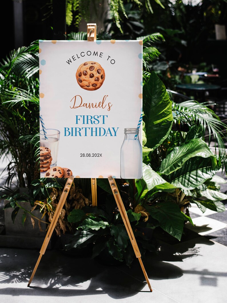 May include: Welcome to Daniel's First Birthday sign with a cookie illustration, a glass of milk and a bottle of milk. The sign is on a wooden easel.