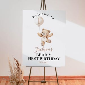 May include: A white rectangular welcome sign on a metal easel featuring a watercolor illustration of a brown teddy bear holding a beige balloon with the number 1. Text reads: Welcome to, Jackson's, BEAR-Y FIRST BIRTHDAY, and the date 25.02.202X.
