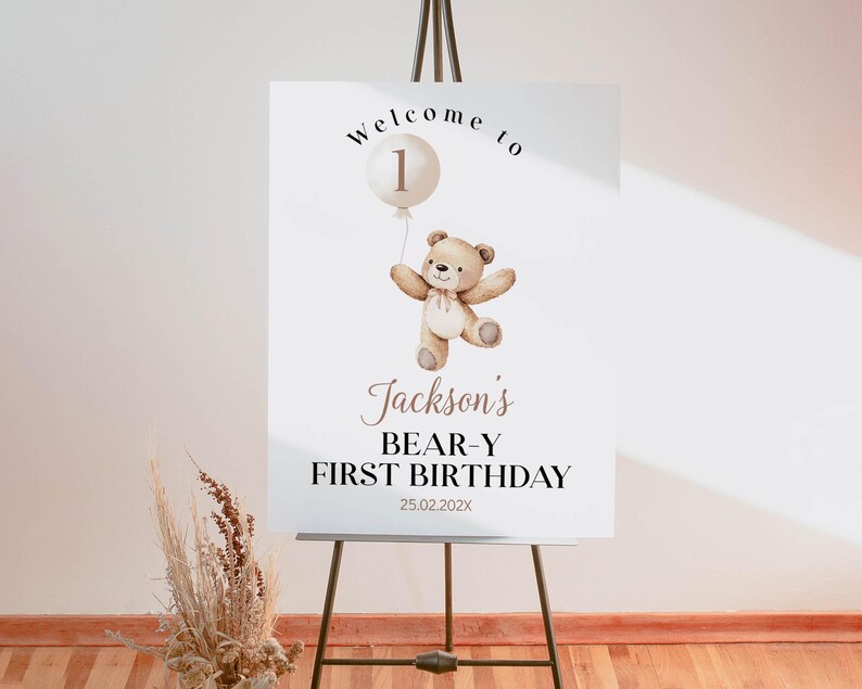 May include: A white rectangular welcome sign on a metal easel featuring a watercolor illustration of a brown teddy bear holding a beige balloon with the number 1. Text reads: Welcome to, Jackson's, BEAR-Y FIRST BIRTHDAY, and the date 25.02.202X.