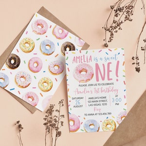 May include: A birthday invitation card and matching envelope liner featuring a watercolor donut pattern with colorful sprinkles. The text reads: AMELIA is a sweet ONE! PLEASE JOIN US TO CELEBRATE Amelia's 1st birthday SATURDAY 26 AUGUST AMELIA'S HOME 123 MAIN STREET, LAS VEGAS. 1235 AT 3:00 PM RSVP TO ANNA AT 123.456.789.