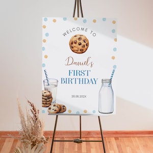 May include: Welcome to Daniel's First Birthday party sign with blue and gold polka dots, a chocolate chip cookie, a glass of milk and a milk bottle. The date is 28.08.202X.