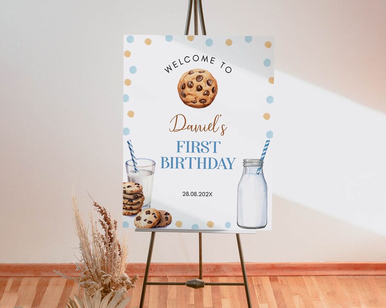 May include: Welcome to Daniel's First Birthday party sign with blue and gold polka dots, a chocolate chip cookie, a glass of milk and a milk bottle. The date is 28.08.202X.