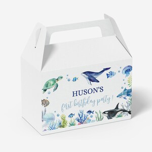 May include: A white cardboard gable box featuring a watercolor ocean theme with a sea turtle, whale, shark, orca, jellyfish, and coral. The text reads HUSON'S first birthday party! in blue script and block lettering on the front panel.