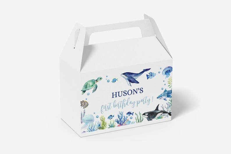 May include: A white cardboard gable box featuring a watercolor ocean theme with a sea turtle, whale, shark, orca, jellyfish, and coral. The text reads HUSON'S first birthday party! in blue script and block lettering on the front panel.