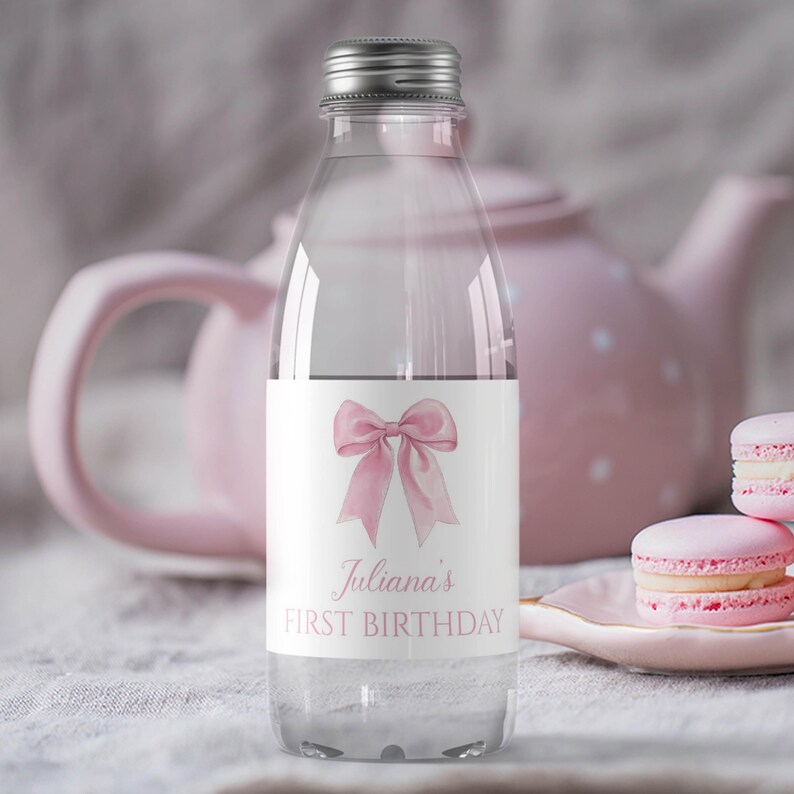Puede incluir: Una botella de agua de pl&aacute;stico transparente con una etiqueta blanca que presenta una ilustraci&oacute;n de un lazo rosa claro y el texto Juliana's FIRST BIRTHDAY en una fuente serif rosa claro. La botella tiene una tapa de rosca plateada.