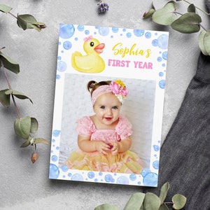 Op de afbeelding: Een gele rubberen eend met een roze strik zit op een witte achtergrond met blauwe aquarel-bellen. De tekst "Sophia's First Year" is geschreven in gele en roze letters. Een foto van een babymeisje in een roze en gele jurk bevindt zich in het midden van de kaart.
