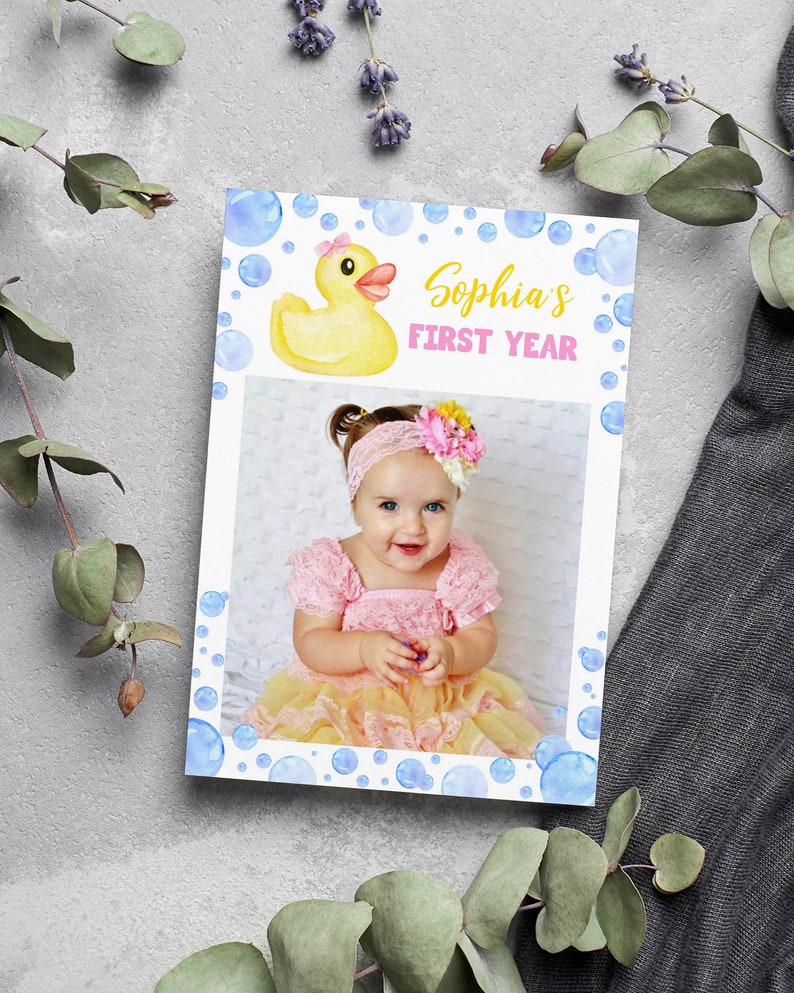Op de afbeelding: Een gele rubberen eend met een roze strik zit op een witte achtergrond met blauwe aquarel-bellen. De tekst "Sophia's First Year" is geschreven in gele en roze letters. Een foto van een babymeisje in een roze en gele jurk bevindt zich in het midden van de kaart.