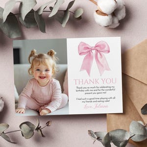 May include: A pink and white thank you card with a watercolor bow illustration. The card features a photo of a smiling baby girl and the text "THANK YOU" with a personal message. Eucalyptus and cotton accents surround the card.