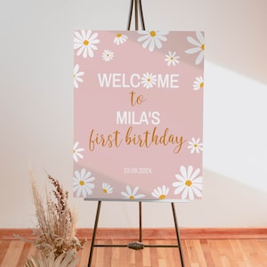Op de afbeelding: Een rechthoekig roze welkomstbord op een zwarte ezel met witte madeliefjesillustraties met gele harten. De tekst luidt WELCOME to MILA'S first birthday 10.08.202X in een combinatie van witte blokletters en gouden cursieve letters.