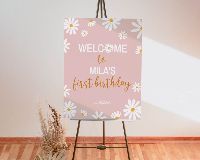Op de afbeelding: Een rechthoekig roze welkomstbord op een zwarte ezel met witte madeliefjesillustraties met gele harten. De tekst luidt WELCOME to MILA'S first birthday 10.08.202X in een combinatie van witte blokletters en gouden cursieve letters.