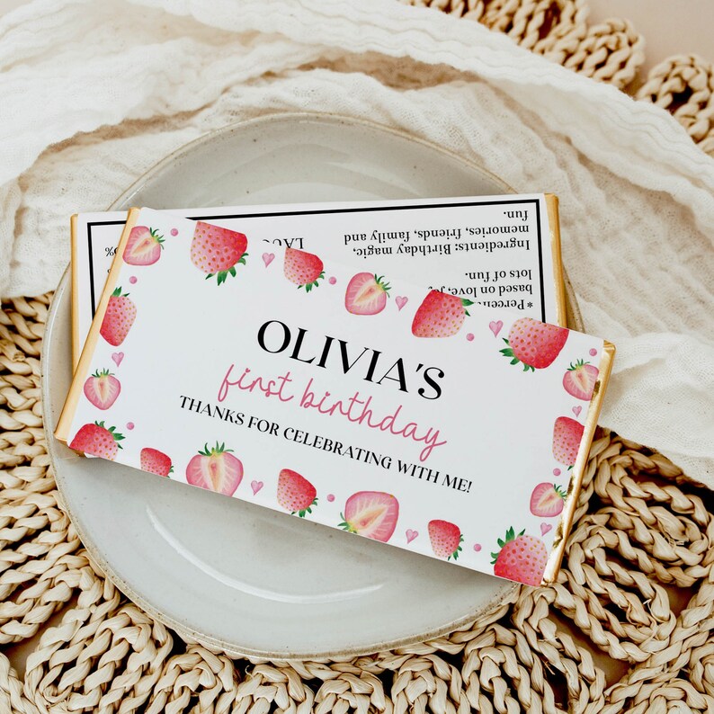 May include: A rectangular chocolate bar wrapper with a white background featuring a watercolor strawberry pattern. The text reads OLIVIA'S first birthday THANKS FOR CELEBRATING WITH ME! and includes a list of ingredients like birthday magic.