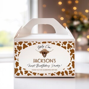 May include: A white cardboard gable box featuring a brown cow print border and a central oval graphic. The design includes an illustration of a Highland cow, the text "Holy Cow", "JACKSON'S First Birthday Party!", and "THANK YOU FOR CELEBRATING WITH ME!"