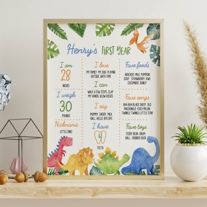 May include: A rectangular framed poster titled Henry's First Year featuring watercolor dinosaur illustrations and tropical leaves. The text lists milestones including 28 inches, 30 pounds, 4 teeth, favorite foods, songs, and toys in colorful fonts.