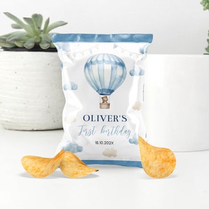 May include: A white chip bag with a blue hot air balloon design, clouds, and a teddy bear. The text "OLIVER'S First birthday" is displayed. Two potato chips are in front of the bag.