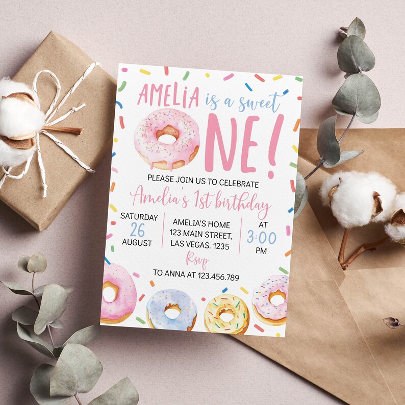 May include: A rectangular birthday invitation card featuring a pink frosted donut and the text AMELIA is a sweet ONE! PLEASE JOIN US TO CELEBRATE Amelia's 1st birthday SATURDAY 26 AUGUST AMELIA'S HOME 123 MAIN STREET, LAS VEGAS. 1235 AT 3:00 PM RSVP TO ANNA AT 123.456.789
