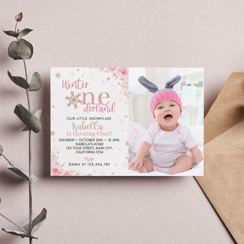 May include: A rectangular birthday invitation card featuring a photo of a baby wearing a pink and grey bunny ear hat. The text reads Winter One-derland, Our Little Snowflake, Isabella is turning One, with event details and a rose and snowflake design.