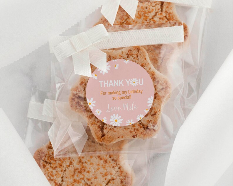 May include: Star-shaped cookies packaged in clear plastic bags, each tied with a white ribbon. A round pink sticker on the front features white daisy illustrations and the text: THANK YOU For making my birthday so special! Love, Mila.