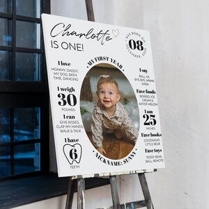 May include: A white milestone birthday sign for a one-year-old named Charlotte, featuring a central oval photo of a baby, text detailing birth date, weight of 30 pounds, height of 25 inches, six teeth, favorite foods, books, toys, and milestones.