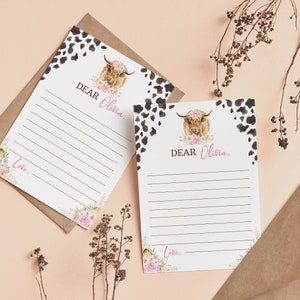 May include: Two blank white cards with black and white cowhide print borders. Each card features a watercolor illustration of a Highland cow with pink flowers. The text "DEAR Olivia." is printed on each card. The cards are on a light brown background.