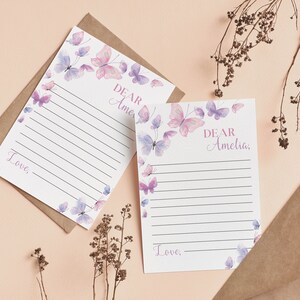 May include: Two sheets of stationery with lined paper and a butterfly design in pink and purple. The text "Dear Amelia" and "Love," are printed on the stationery. The stationery is on a light pink surface with a brown envelope.