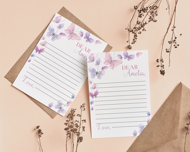 May include: Two sheets of stationery with lined paper and a butterfly design in pink and purple. The text "Dear Amelia" and "Love," are printed on the stationery. The stationery is on a light pink surface with a brown envelope.