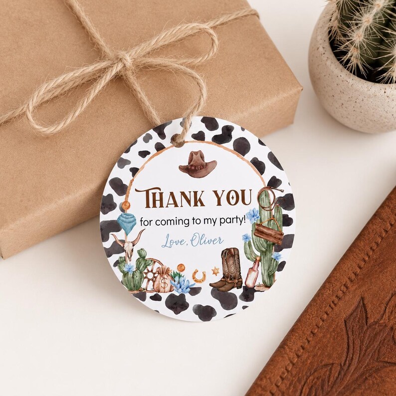 May include: A round paper gift tag with a black and white cow print border. The center features illustrations of a brown cowboy hat, a cactus, a lasso, a boot, and a skull. Text reads: Thank you for coming to my party! Love, Oliver.