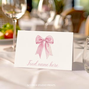 May include: A white rectangular folded place card featuring a centered illustration of a light pink ribbon bow. Below the bow, the text "Food name here" is printed in a cursive pink font. The card is positioned on a white fabric surface.
