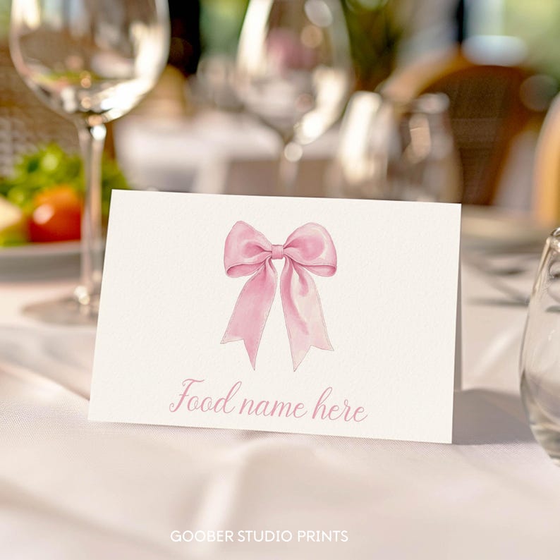 May include: A white rectangular folded place card featuring a centered illustration of a light pink ribbon bow. Below the bow, the text "Food name here" is printed in a cursive pink font. The card is positioned on a white fabric surface.