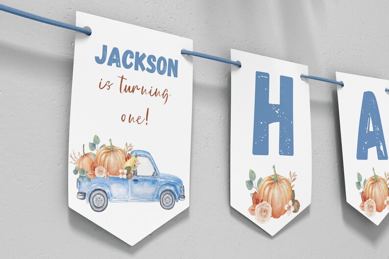 May include: A white decorative banner featuring blue text that reads JACKSON is turning one! and a blue truck illustration carrying orange pumpkins and flowers. The banner includes additional panels with large blue letters and pumpkin graphics.