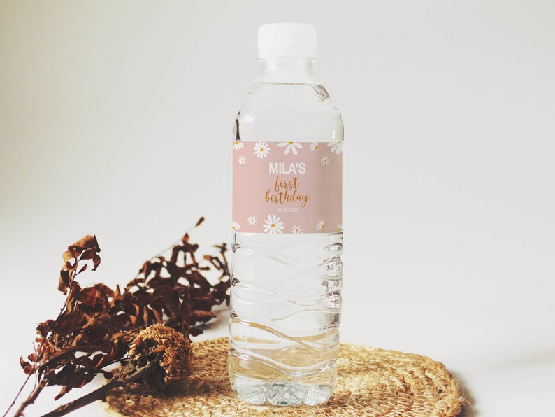 May include: A clear plastic water bottle with a white cap and a custom label. The label features a dusty pink background with white daisy illustrations and the text "MILA'S first birthday 10.08.202X" printed in white and gold-colored script.