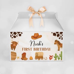 Puede incluir: Una caja blanca con un lazo de sat&eacute;n beige en la parte superior. El frente presenta una ilustraci&oacute;n de tem&aacute;tica western con un sombrero de vaquero marr&oacute;n, un toro, un caballo, un barril, botas, una fogata, una carreta, un cactus y el texto Noah's FIRST BIRTHDAY.