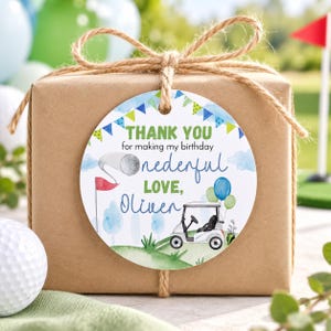 May include: A round white gift tag tied to a brown paper package with twine. The tag features a golf theme with a golf cart, a golf ball on a tee, and blue and green bunting. Text reads: THANK YOU for making my birthday onederful LOVE, Oliver.