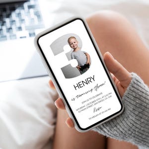 Op de afbeelding: Een digitale verjaardagsuitnodiging op een smartphonescherm met een groot cijfer drie waarin een foto van een jonge jongen is verwerkt. De tekst luidt HENRY is turning three! JOIN US TO CELEBRATE SATURDAY OCTOBER 15TH AT 2:00 PM 123 MAIN STREET SAN DIEGO CA 1345 RSVP TO MEGAN AT 123.456.789.
