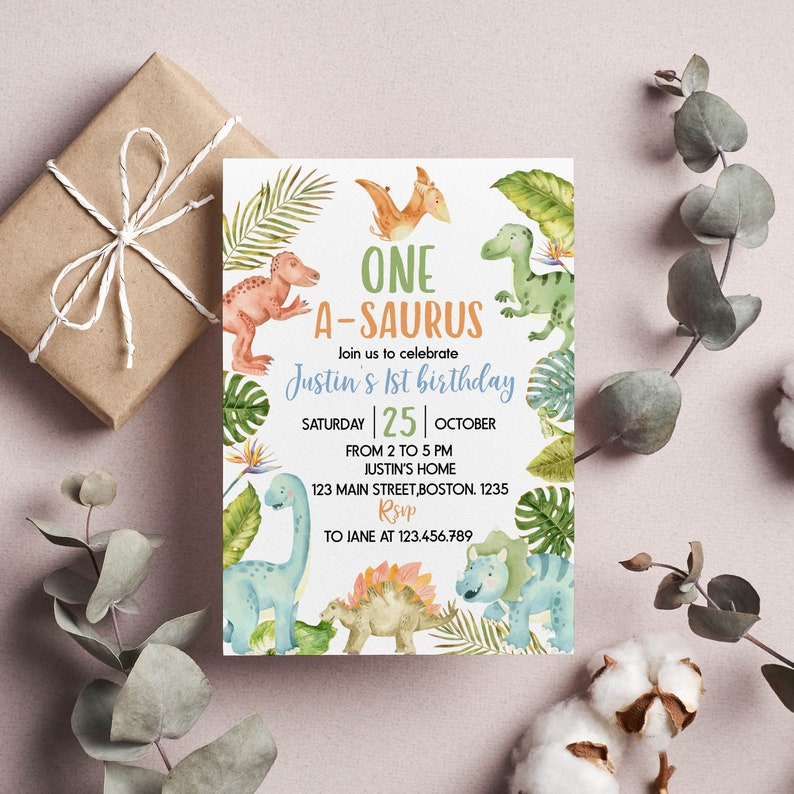 Op de afbeelding: Een rechthoekige verjaardagsuitnodigingskaart met aquarelillustraties van diverse dinosaurussen en tropische bladeren. De tekst luidt: ONE A-SAURUS, Join us to celebrate Justin's 1st birthday, SATURDAY 25 OCTOBER, FROM 2 TO 5 PM, JUSTIN'S HOME, 123 MAIN STREET, BOSTON, 1235, RSVP TO JANE AT 123.456.789.