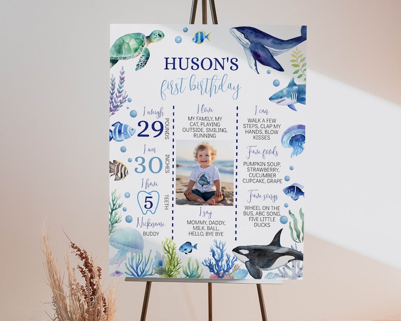 May include: A blue and white first birthday milestone poster with a watercolor ocean theme. The poster features a photo of a baby boy, along with details about the baby's age, weight, height, and favorite foods and songs. The poster also includes a variety of ocean-themed illustrations, such as fish, sea turtles, and a whale.