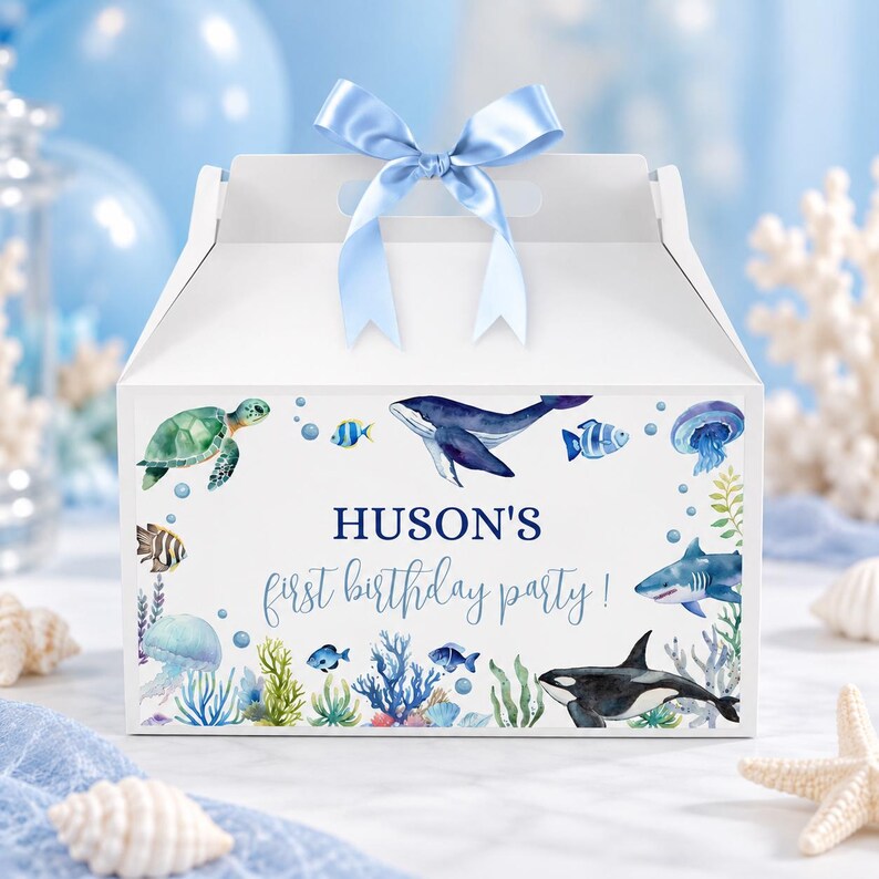May include: A white cardboard gable box with a light blue satin ribbon bow on top. The front features watercolor illustrations of sea creatures including a turtle, whale, shark, orca, and jellyfish, with the text HUSON'S first birthday party!