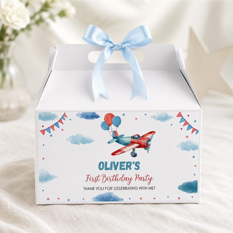 May include: A white cardboard gable box with a light blue satin ribbon bow on top. The front features a watercolor illustration of a red and blue airplane with balloons, blue clouds, and red and blue bunting. Text reads: OLIVER'S First Birthday Party THANK YOU FOR CELEBRATING WITH ME!
