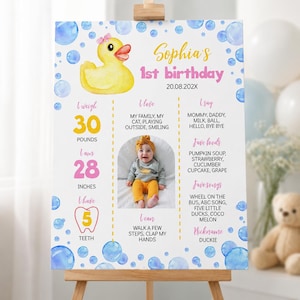 May include: A birthday milestone sign featuring a yellow rubber duck with a pink bow, blue bubbles, and text including Sophia's 1st birthday, 20.08.202X, 30 pounds, 28 inches, 5 teeth, and personal details alongside a central photograph of a child.