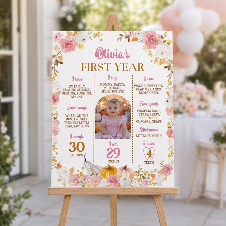 May include: A white milestone birthday sign for Olivia's first year, decorated with pink and white roses and orange pumpkins. It features text detailing favorite songs, foods, and milestones, along with a central photo of a baby in a pink dress.
