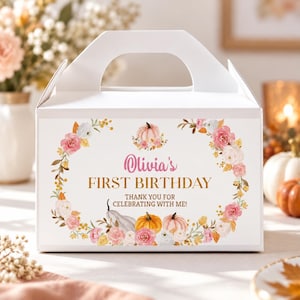 May include: A white cardboard gable box featuring a floral wreath design with pink and white flowers, orange and white pumpkins, and the text "Olivia's FIRST BIRTHDAY THANK YOU FOR CELEBRATING WITH ME!" printed in pink and gold on the front panel.