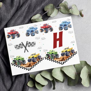May include: Monster truck birthday banner with the letter H in red. The banner features a checkered flag and several monster trucks in various colors, including red, blue, green, and yellow.