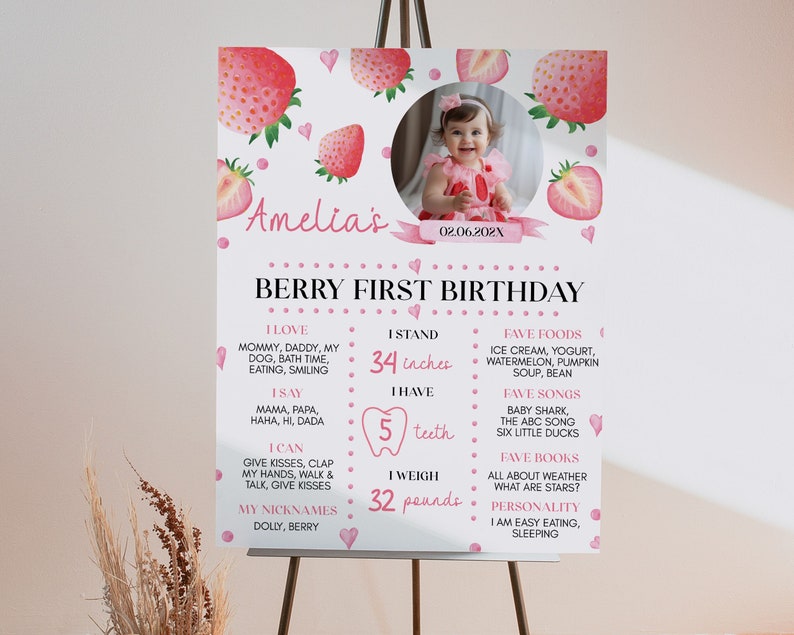 Op de afbeelding: Een wit verjaardagsbord met aquarel aardbeien, roze hartjes en een ronde kinderfoto. De tekst bevat Amelia's Berry First Birthday, 86 cm, 5 tanden, 14,5 kg, en lijsten met favoriete etenswaren, liedjes, boeken en persoonlijkheidskenmerken.