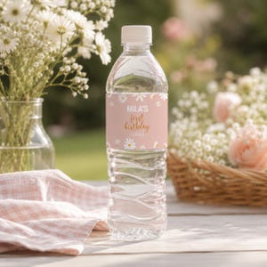 May include: A clear plastic water bottle with a white cap and a custom label. The label is light pink with a white daisy pattern and features the text "MILA'S first birthday" in white and gold script lettering centered on the front.