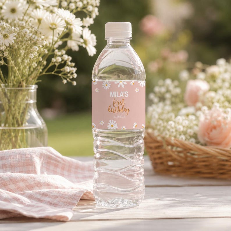 May include: A clear plastic water bottle with a white cap and a custom label. The label is light pink with a white daisy pattern and features the text "MILA'S first birthday" in white and gold script lettering centered on the front.