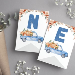 May include: Two white rectangular pennant flags featuring a large blue letter N or E at the top. Below each letter is a watercolour illustration of a blue pickup truck carrying orange pumpkins and autumn flowers, with floral accents at the top.