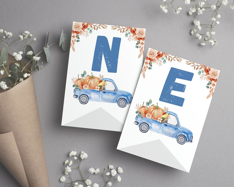 May include: Two white rectangular pennant flags featuring a large blue letter N or E at the top. Below each letter is a watercolour illustration of a blue pickup truck carrying orange pumpkins and autumn flowers, with floral accents at the top.